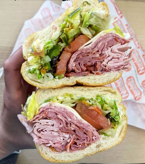 A hand holding a cross-section of a sandwich filled with layers of deli meats, lettuce, and tomato on soft bread.