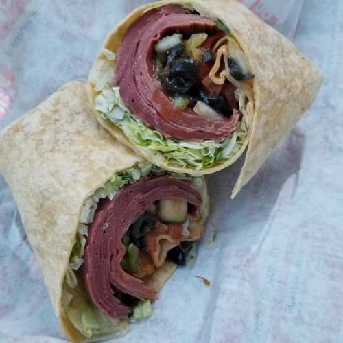 A close-up of a sliced wrap filled with layers of deli meat, lettuce, tomatoes, and olives, wrapped in a tortilla.