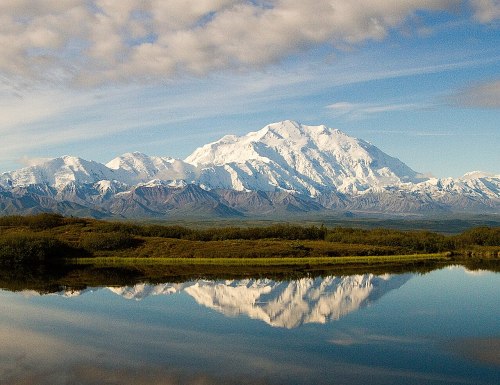 Snow-capped mountains reflect in a calm lake under a blue sky with scattered clouds.