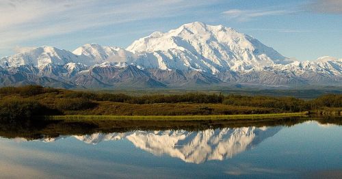 Snow-capped mountains reflect in a calm lake under a clear blue sky, surrounded by lush greenery.