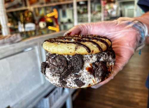 A hand holding a cookie sandwich filled with cookies and cream ice cream, drizzled with chocolate sauce.