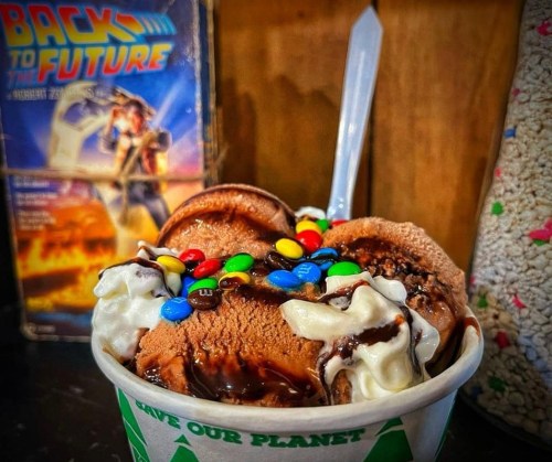 A bowl of chocolate ice cream topped with whipped cream, chocolate syrup, and colorful candy, with a "Back to the Future" box in the background.