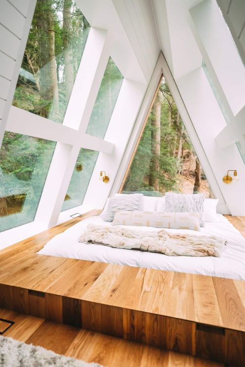 Cozy bedroom with large triangular windows, wooden flooring, and a plush bed surrounded by lush greenery.