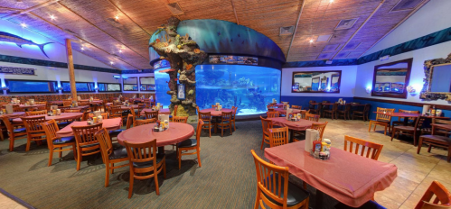 Interior of a restaurant featuring wooden tables, chairs, and a large aquarium as a centerpiece, with blue lighting.