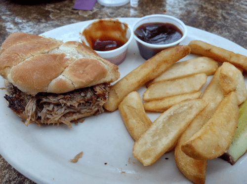 A plate with a pulled pork sandwich, two dipping sauces, and a side of golden fries.