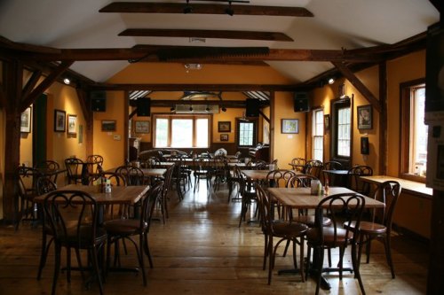Cozy restaurant interior with wooden beams, empty tables, and warm orange walls, featuring artwork and natural light.