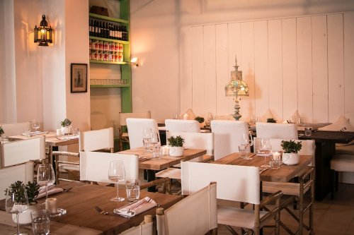 A cozy restaurant interior with wooden tables, white chairs, and potted plants, featuring soft lighting and shelves of bottles.