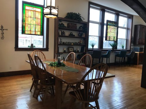 A spacious dining area with a wooden table, chairs, and stained glass windows, featuring shelves and a kitchen in the background.