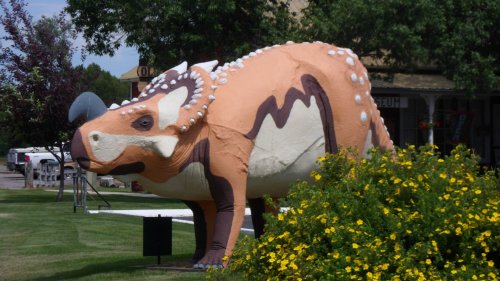 A large, colorful dinosaur sculpture stands in a grassy area, surrounded by yellow flowers and trees.