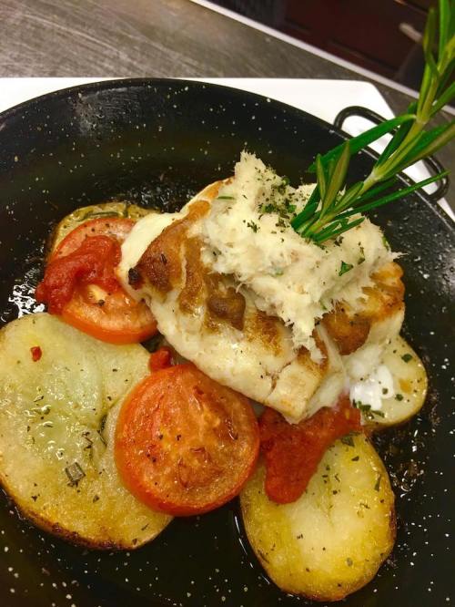 A plated dish featuring fish topped with crab, served on a bed of sliced potatoes and tomatoes, garnished with rosemary.