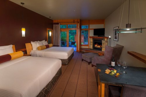 Cozy hotel room with two beds, a fireplace, and a view of nature, featuring a table with wine and snacks.