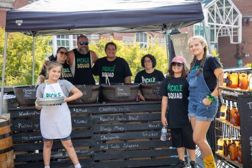 A group of people in "Pickle Squad" shirts at a market booth, showcasing various pickles and products.