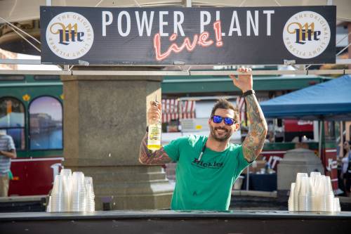 A bartender smiles and holds a drink at a lively outdoor event with a "Power Plant Live!" sign above.
