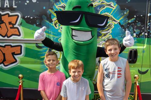 Three smiling children pose in front of a large green mascot with sunglasses, set against a colorful backdrop.