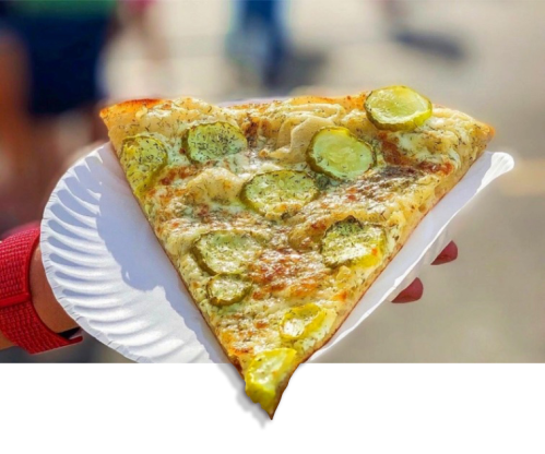 A slice of pizza topped with cheese and sliced pickles, held on a paper plate against a blurred background.