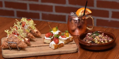 A wooden platter with crispy tacos, bruschetta topped with tomatoes, and a bowl of savory dish, alongside a cocktail.