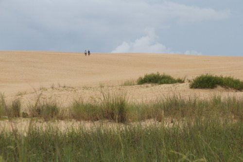 Two people walking on a sandy dune, with green grass in the foreground and a cloudy sky above.