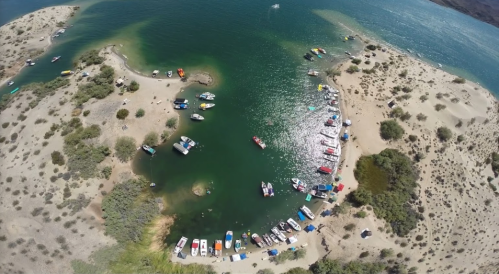Aerial view of a sandy cove with boats anchored in clear green water, surrounded by sparse vegetation and hills.