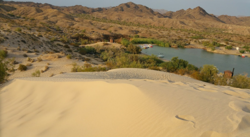 A sandy dune overlooks a river surrounded by lush greenery and mountains in the background.