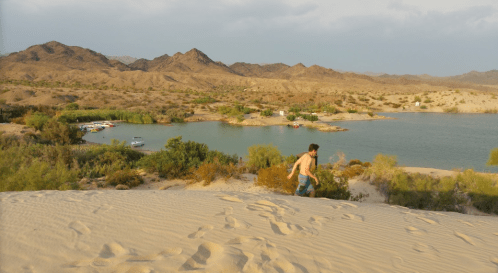 A person climbs a sandy dune overlooking a river and mountains in a desert landscape.