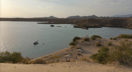 A serene river scene with a boat, sandy shore, and distant mountains under a partly cloudy sky.