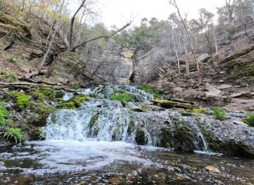A serene waterfall cascades over rocky terrain, surrounded by trees and greenery in a tranquil natural setting.
