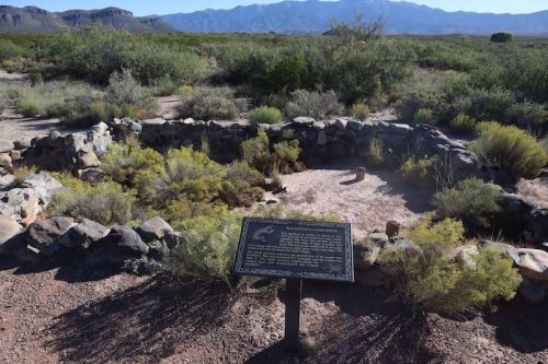 A stone circle surrounded by shrubs, with a plaque detailing its significance, set against a mountain backdrop.