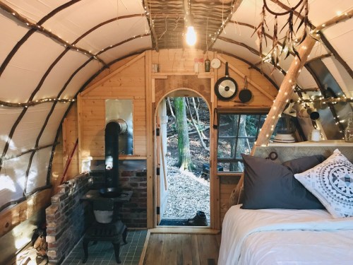 Cozy interior of a tiny cabin with a bed, wood stove, and rustic decor, surrounded by trees and soft lighting.