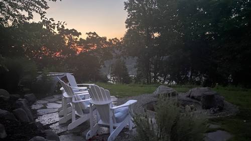 A serene outdoor scene at dusk with white chairs facing a fire pit, surrounded by trees and a view of the sunset.