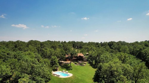 Aerial view of a house surrounded by trees, featuring a swimming pool and a spacious green lawn under a clear blue sky.