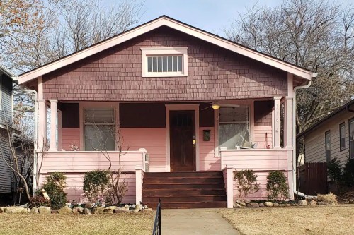 A charming pink house with a front porch, surrounded by greenery and trees, on a clear day.