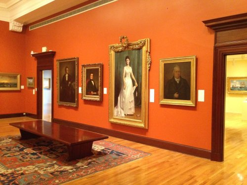 A gallery with orange walls displaying several framed portraits and a wooden table in the center.