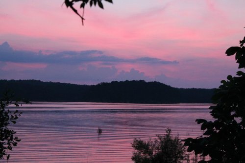 A serene lake at sunset, reflecting pink and purple hues in the sky, surrounded by trees and gentle ripples on the water.