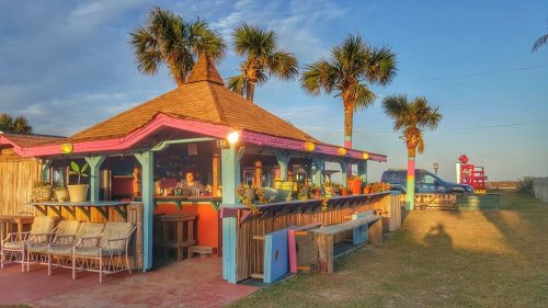 A colorful beach bar with palm trees, outdoor seating, and a sunset sky in the background.
