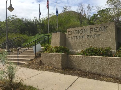 Sign for Ensign Peak Nature Park with stairs leading up, surrounded by greenery and flags in the background.