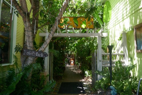 A lush, green pathway leads through an archway with a sign, surrounded by tropical plants and trees.