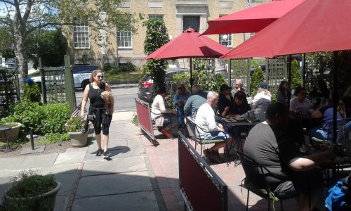 A busy outdoor café with patrons dining under red umbrellas, while a woman walks by with a small dog.