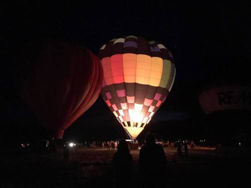 Colorful hot air balloons illuminated at night, with silhouettes of people watching in the foreground.