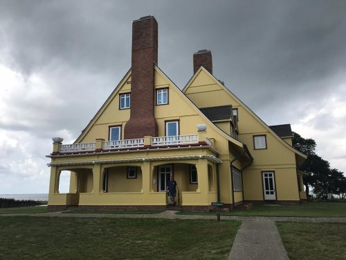 A large yellow house with two brick chimneys, set against a cloudy sky, with a person standing on the porch.
