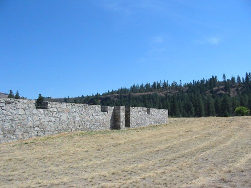 Stone wall ruins in a grassy field, surrounded by trees under a clear blue sky.