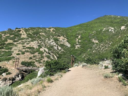 A dirt path leads to a bridge over a rocky area, surrounded by lush green hills under a clear blue sky.