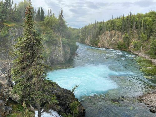 A serene river flows through a lush, green landscape, surrounded by tall trees and rocky cliffs under a cloudy sky.