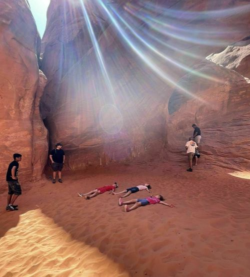 A group of people in a sandy canyon, some lying down while others explore the rocky surroundings under bright sunlight.