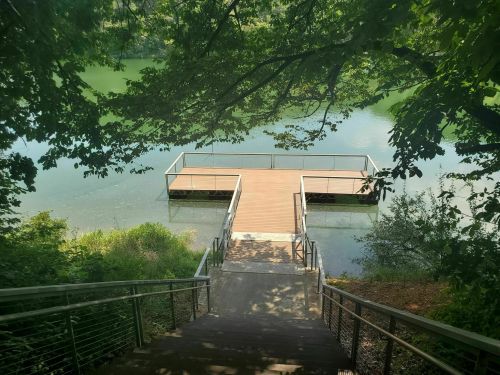 A wooden dock extends into a calm green lake, surrounded by lush trees and foliage. Steps lead down to the water.