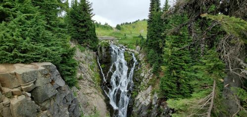 A cascading waterfall surrounded by lush green trees and rocky cliffs, set against a cloudy sky.