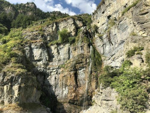A rocky cliff with greenery and a small waterfall cascading down the side under a blue sky with scattered clouds.