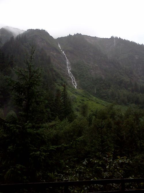 A misty mountain landscape with a waterfall cascading down lush green slopes, surrounded by dense trees.