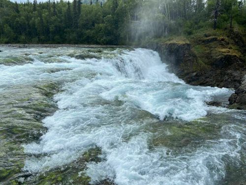 A rushing waterfall cascades over rocky terrain, surrounded by lush green trees and mist rising from the water.