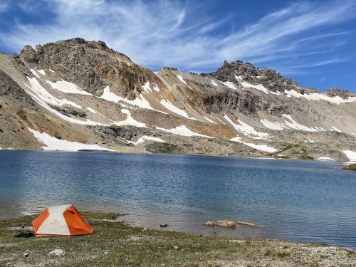 A bright orange tent by a serene lake, surrounded by rugged mountains and patches of snow under a blue sky.