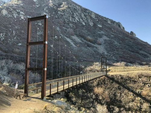 A suspension bridge spans a valley, surrounded by rocky hills and sparse vegetation under a clear blue sky.